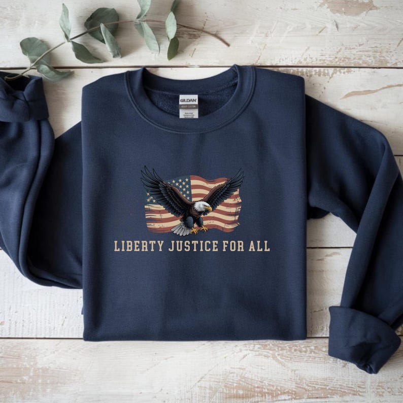 May include: Navy blue sweatshirt featuring an eagle with outstretched wings in front of a distressed American flag. The text "LIBERTY JUSTICE FOR ALL" is printed below the graphic. The sweatshirt has a crew neck and long sleeves.