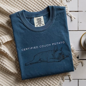May include: A blue Comfort Colors t-shirt with the words "CERTIFIED COUCH POTATO" above a black illustration of a French Bulldog. The shirt is folded on a white surface.