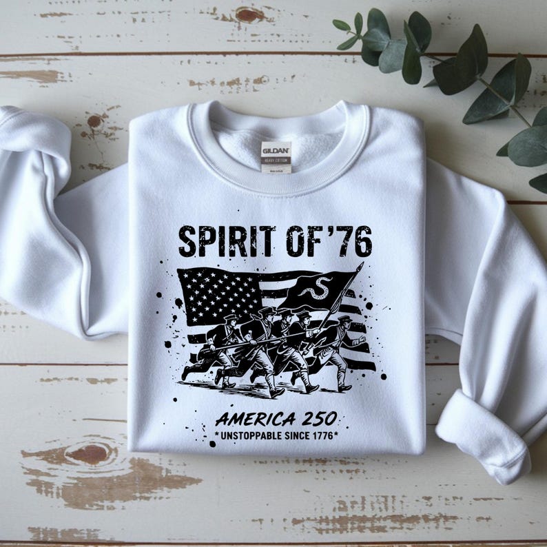May include: White sweatshirt with a graphic design featuring the text "SPIRIT OF '76" above an illustration of soldiers carrying a flag. The American flag is in the background, with the text "AMERICA 250 *UNSTOPPABLE SINCE 1776*" below.