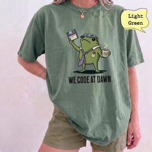 May include: A light green t-shirt featuring a graphic of a frog wearing a visor and tie, holding a floppy disk and a coffee mug. The text "WE CODE AT DAWN" is printed below the frog. The shirt has a crew neckline and short sleeves.