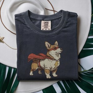 May include: A charcoal gray short-sleeve t-shirt featuring a graphic of a Corgi dog dressed as a superhero. The dog wears a red cape, a red eye mask, and a cream-colored shirt with a yellow lightning bolt emblem. The inner neck label reads Comfort Colors.