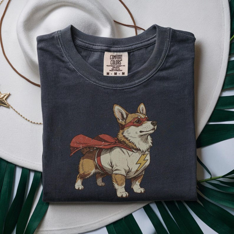 May include: A charcoal gray short-sleeve t-shirt featuring a graphic of a Corgi dog dressed as a superhero. The dog wears a red cape, a red eye mask, and a cream-colored shirt with a yellow lightning bolt emblem. The inner neck label reads Comfort Colors.
