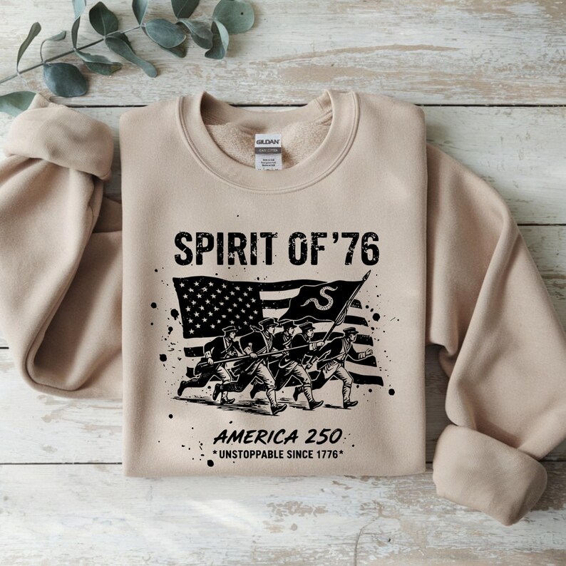 May include: A tan crewneck sweatshirt with the text "SPIRIT OF '76" above a black and white graphic of soldiers carrying a flag. Below the graphic is the text "AMERICA 250 *UNSTOPPABLE SINCE 1776*".