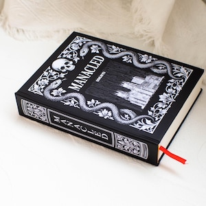 May include: A hardback book with a black cover, featuring the title "MANACLED" in white. The design includes snakes, a skull, and floral patterns. A red bookmark is inserted.