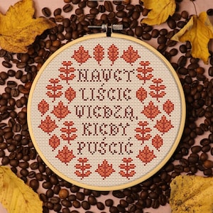 May include: A cross-stitch embroidery hoop with the text "NAWET LIŚCIE WIEDZĄ, KIEDY PUŚCIĆ" in brown thread, surrounded by fall leaves. The hoop is set against a backdrop of coffee beans and autumn leaves.