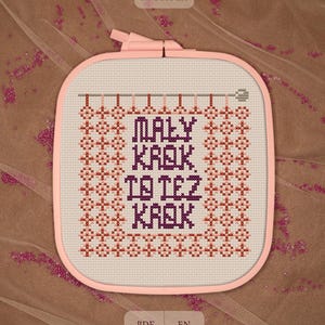 May include: A cross-stitch embroidery in a pink square frame. The design features the text "MAŁY KROK TEŻ KROK" in purple, surrounded by a border of red and white cross-stitch patterns. The top of the frame has the text "whisper of the stitch".