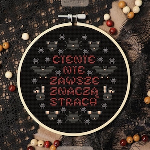 May include: A black cross-stitch embroidery hoop with the text "CIENIE NIE ZAWSZE ZNACZA STRACH" in red. The design includes bats, spiders, and stars. The hoop is set against a black lace background with wooden beads.