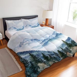 Könnte beinhalten: Ein Bett mit Bettbezug und zwei Kissenbezügen mit einem Aquarell-Berglandschaftsdesign. Die Bettwäsche hat Blau- und Grüntöne, mit einem Wald am unteren Rand und Bergen im Hintergrund.