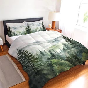 May include: A bedding set featuring a duvet cover and two pillowcases with a watercolour print of a misty forest landscape. The design displays layers of dark green pine trees fading into soft grey and white mountains in the background.