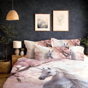 Può includere: Un letto con un set copripiumino a tema unicorno rosa e bianco. La biancheria da letto presenta un grande unicorno bianco con una criniera fluente e un corno, sullo sfondo di fiori di ciliegio rosa. Il set include federe e un copripiumino.