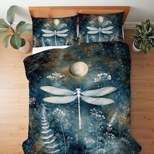 May include: A bedding set featuring a dark blue and brown watercolor background with a large white dragonfly, a full moon, and white botanical silhouettes. The design includes a duvet cover and two matching pillowcases with a consistent pattern.