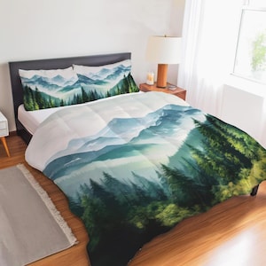 May include: A bedding set featuring a comforter and two pillowcases with a printed landscape design. The pattern displays a misty mountain range in shades of blue and white, layered behind a dense forest of dark green pine trees.