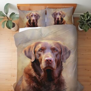 Puede incluir: Un juego de cama que incluye una funda nórdica y dos fundas de almohada a juego con un retrato detallado de un Labrador Retriever marrón. El perro tiene ojos color café y un pelaje texturizado sobre un fondo de tonos neutros y suaves.