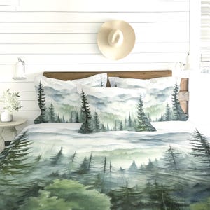 May include: A bedding set featuring a watercolour-style print of a misty forest with evergreen trees in shades of green, grey, and white. The set includes a quilt and two matching pillow shams, all displaying a continuous landscape of pine trees.