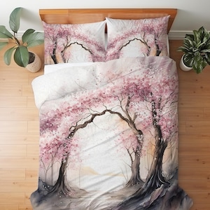 以下が含まれることがあります： ピンク、ブラウン、ホワイトの色合いで桜の木が描かれた水彩画風プリントの掛け布団カバーと枕カバー2枚の寝具セット。満開の木々が並ぶ小道が描かれており、生地全体にアーチのような効果を生み出しています。