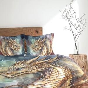 Op de afbeelding: Een beddengoedset met een aquarel-stijl illustratie van een gouden draak met blauwe ogen. Het ontwerp bevat twee vierkante kussenslopen en een groot dekbedovertrek, die allemaal hetzelfde gedetailleerde drakenkopmotief op een donkere achtergrond tonen.