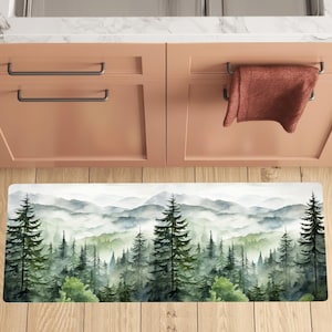 May include: A watercolor-style kitchen rug with a misty mountain scene featuring a forest of green trees. The rug is rectangular and has a white background.