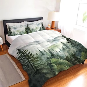 May include: A bedding set featuring a watercolor-style print of a misty forest with dark green pine trees and soft gray mountains. The design covers the comforter and two matching pillowcases, displaying a continuous landscape of trees.