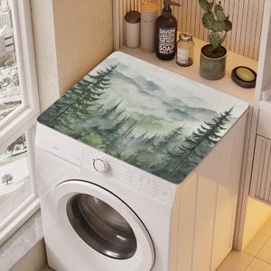 May include: A white washing machine with a watercolor-style forest scene printed on the top. The scene features a misty forest with green trees and a mountain range in the background.