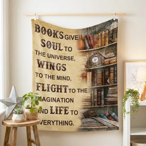Op de afbeelding: Een beige wandtapijt met de quote "BOOKS GIVE SOUL TO THE UNIVERSE, WINGS TO THE MIND, FLIGHT TO THE IMAGINATION AND LIFE TO EVERYTHING." De rechterkant toont een boekenplank illustratie met boeken en een klok.