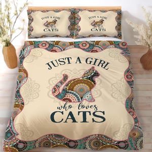 Może przedstawiać: Beżowy zestaw pościeli z dwoma poszewkami i kołdrą. Kołdra i poszewki mają napis "JUST A GIRL who loves CATS" i dekoracyjny wzór kota. Zestaw ma kolorową obwódkę w stylu mandali.