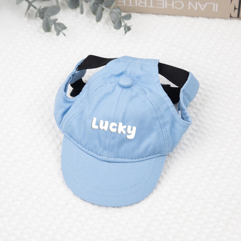 May include: A light blue baseball cap for a pet, featuring the word "Lucky" in white lettering. The cap has a curved brim and a black strap for securing it. The cap is made of a soft material.
