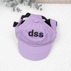 May include: A lavender baseball cap for a pet, featuring the embroidered letters "dss" in black. The cap has a black strap and is designed to fit over the pet's head, offering a stylish accessory.