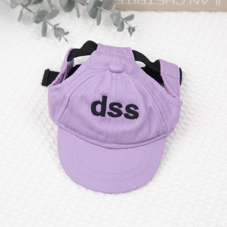 May include: A lavender baseball cap for a pet, featuring the embroidered letters "dss" in black. The cap has a black strap and is designed to fit over the pet's head, offering a stylish accessory.