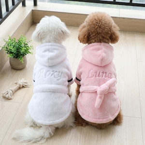 May include: Two small dogs wearing matching hooded robes. One dog is wearing a white robe with "Roxy" embroidered on the back, and the other is wearing a pink robe with "Luna" embroidered on the back. Both robes have a belt.
