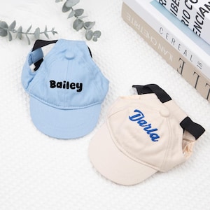 May include: Two pet baseball caps. One is light blue with the name "Bailey" in black. The other is beige with "Darla" in blue. Both have a black strap and a button on top. The caps are designed for pets.