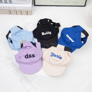 May include: Five baseball caps for pets in various colors: light blue, black, blue, lavender, and beige. Each cap has a curved brim and a strap. The caps are embroidered with names: "Ellie", "Buddy", "Cooper", "dss", and "Darla".