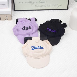 May include: Three pet baseball caps in lavender, black, and beige. Each cap has a curved brim and adjustable straps. The lavender cap has "dss" embroidered in black, the black cap has "Rosie" in purple, and the beige cap has "Darla" in blue.