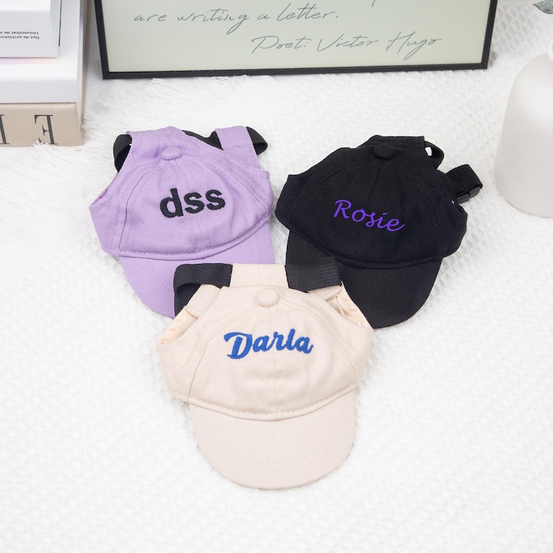May include: Three pet baseball caps in lavender, black, and beige. Each cap has a curved brim and adjustable straps. The lavender cap has "dss" embroidered in black, the black cap has "Rosie" in purple, and the beige cap has "Darla" in blue.