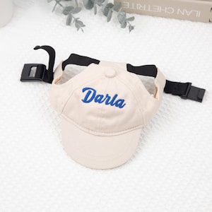 May include: A beige baseball cap with the name "Darla" embroidered in blue. The cap has a black adjustable strap and buckle. The hat is designed for a pet.