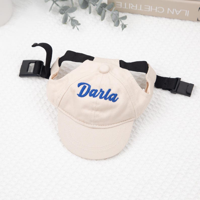 May include: A beige baseball cap with the name "Darla" embroidered in blue. The cap has a black adjustable strap and buckle. The hat is designed for a pet.