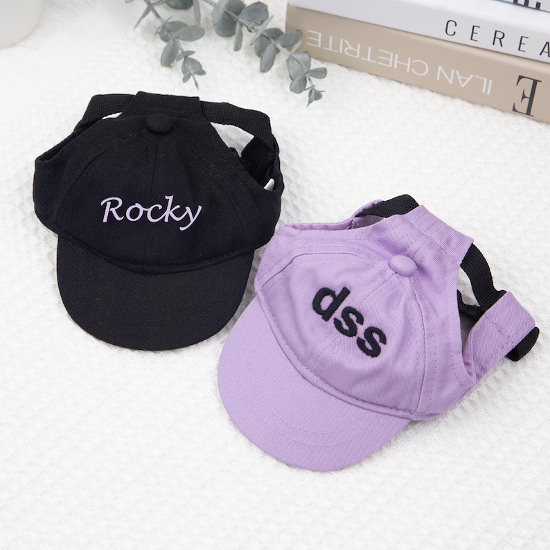 May include: Two baseball caps for pets. One is black with "Rocky" embroidered in white, and the other is lavender with "dss" embroidered in black. Both have adjustable straps.