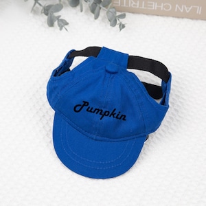 May include: A royal blue baseball cap for pets, featuring the embroidered word "Pumpkin" in black script. The cap has a black strap for securing it to the pet's head.