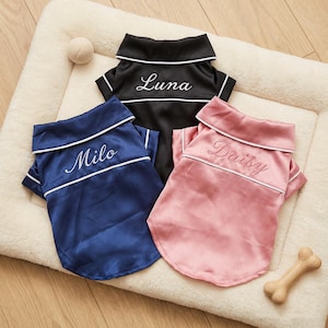 May include: Three satin dog pyjamas in navy blue, black, and pink. Each has a white trim and a name embroidered on the chest: "Milo", "Luna", and "Daisy". A dog bone and a ball are also in the image.