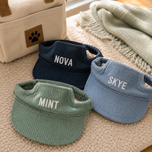 May include: Three corduroy pet visors in navy blue, light blue, and sage green. Each visor features an elasticated band and white embroidered text on the front brim reading NOVA, SKYE, and MINT respectively, designed for pet headwear.
