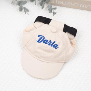 May include: A beige baseball cap with the name "Darla" embroidered in blue script. The cap has a curved brim and a black strap. The cap is designed for a pet.