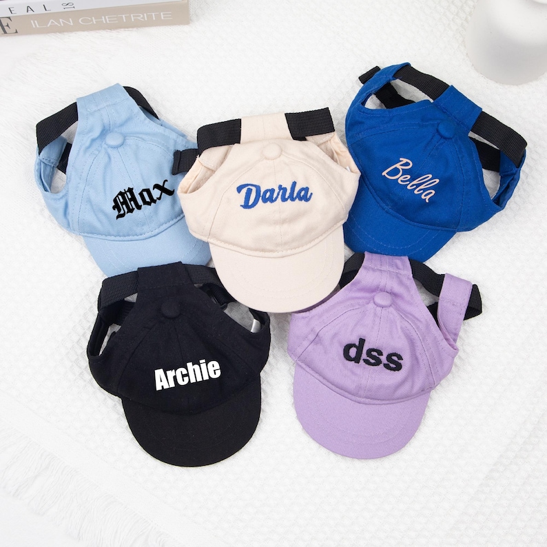 May include: A collection of five pet baseball caps in various colors: light blue, beige, royal blue, black, and lavender. Each cap features a different name embroidered on the front in a contrasting color. The caps have a black strap for securing to the pet's head.