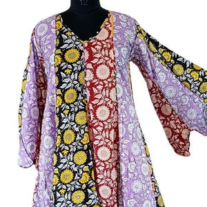 Könnte beinhalten: Ein langes, fließendes Kleid mit V-Ausschnitt und langen Ärmeln. Das Kleid hat Stoffbahnen in verschiedenen Blumenmustern, darunter Sonnenblumen, und Farbtönen von Schwarz, Gelb, Rot und Lila. Das Kleid wird auf einer Schaufensterpuppe präsentiert.