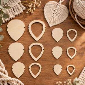 May include: A collection of light-colored wooden teardrop-shaped cutouts arranged in three rows. The set includes solid teardrop shapes and open teardrop frames in various sizes, each featuring small notches on the sides for crafting projects.