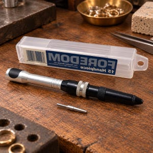 May include: A Foredom 15 handpiece tool with a silver and black cylindrical body, featuring a knurled grip and a metallic finish. The tool is positioned in front of its clear plastic storage case, which displays the brand name and product text.