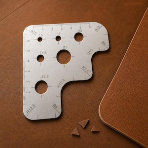 Può includere: Uno strumento in metallo argentato con fori di varie dimensioni e misure in centimetri. Lo strumento ha un bordo righello e bordi curvi con marcature come R10 e R12.5. Lo strumento è su una superficie in pelle marrone.