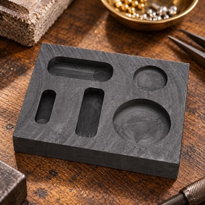 May include: A rectangular dark grey charcoal casting block featuring multiple recessed cavities of varying shapes and sizes, including two elongated slots, one smaller horizontal slot, one small circular depression, and one larger circular depression.