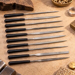May include: A set of ten metal needle files, each featuring a smooth, cylindrical black handle and a distinctively shaped steel blade, including round, flat, and tapered profiles, arranged in a parallel formation for precision metalwork.