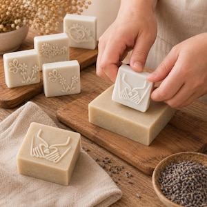May include: A collection of square, light beige soap bars featuring embossed designs including hands forming a heart shape and floral patterns. One soap bar is being pressed with a square stamp featuring a heart-shaped hand design.