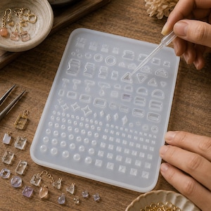 Puede incluir: Un molde de silicona translúcido rectangular que presenta una variedad de pequeñas cavidades geométricas para moldear formas de resina, incluyendo círculos, cuadrados, triángulos, estrellas y varios bucles, acompañado de un gotero de plástico.