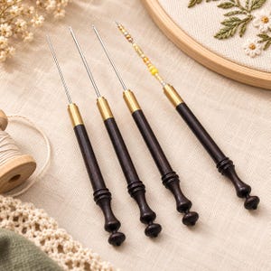May include: Four crochet hooks with dark brown wooden handles and brass ferrules. Three hooks have plain silver metal tips, while the fourth hook features a series of small yellow, white, and metallic beads threaded onto the thin metal shaft.